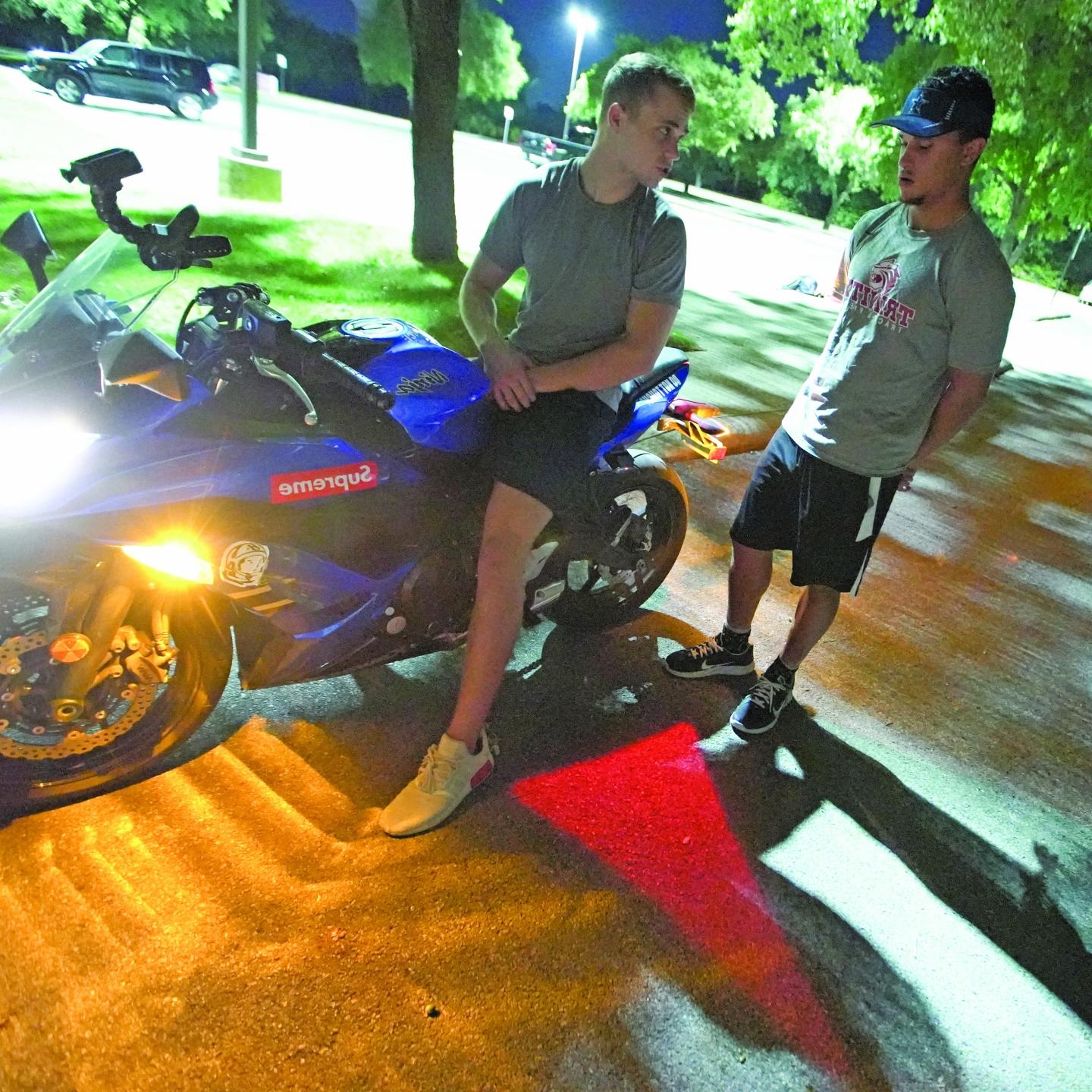 Chris Stewart and Bobby Magee sit on a motorcycle with a red turn signal projected on the asphalt below them.